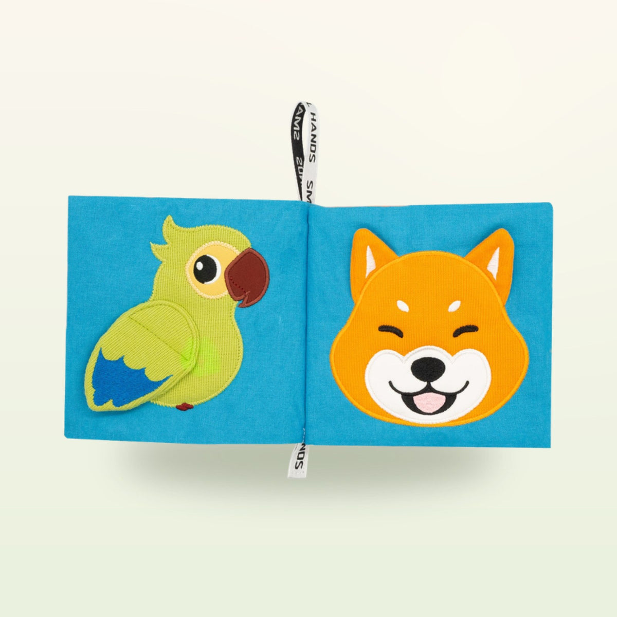 Small Hands Pets Montessori cloth book open – green parrot and smiling Shiba dog on a soft cream background – from 0 months