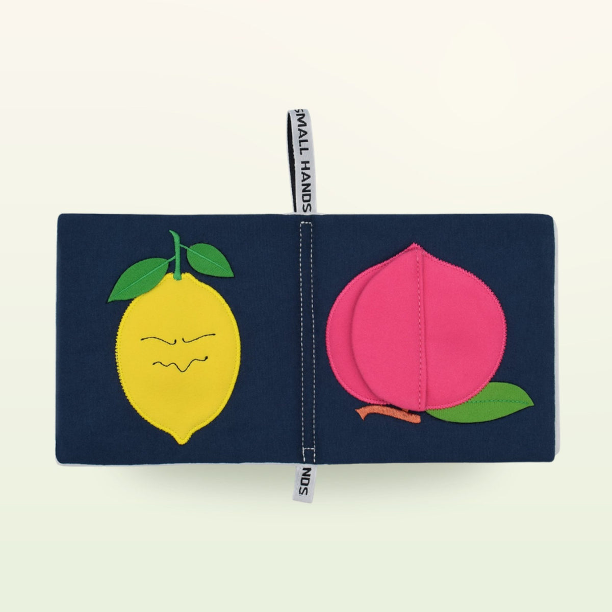 Small Hands Colorful Fruits Montessori cloth book open – yellow lemon with a face and pink peach on a soft cream background – from 0 months