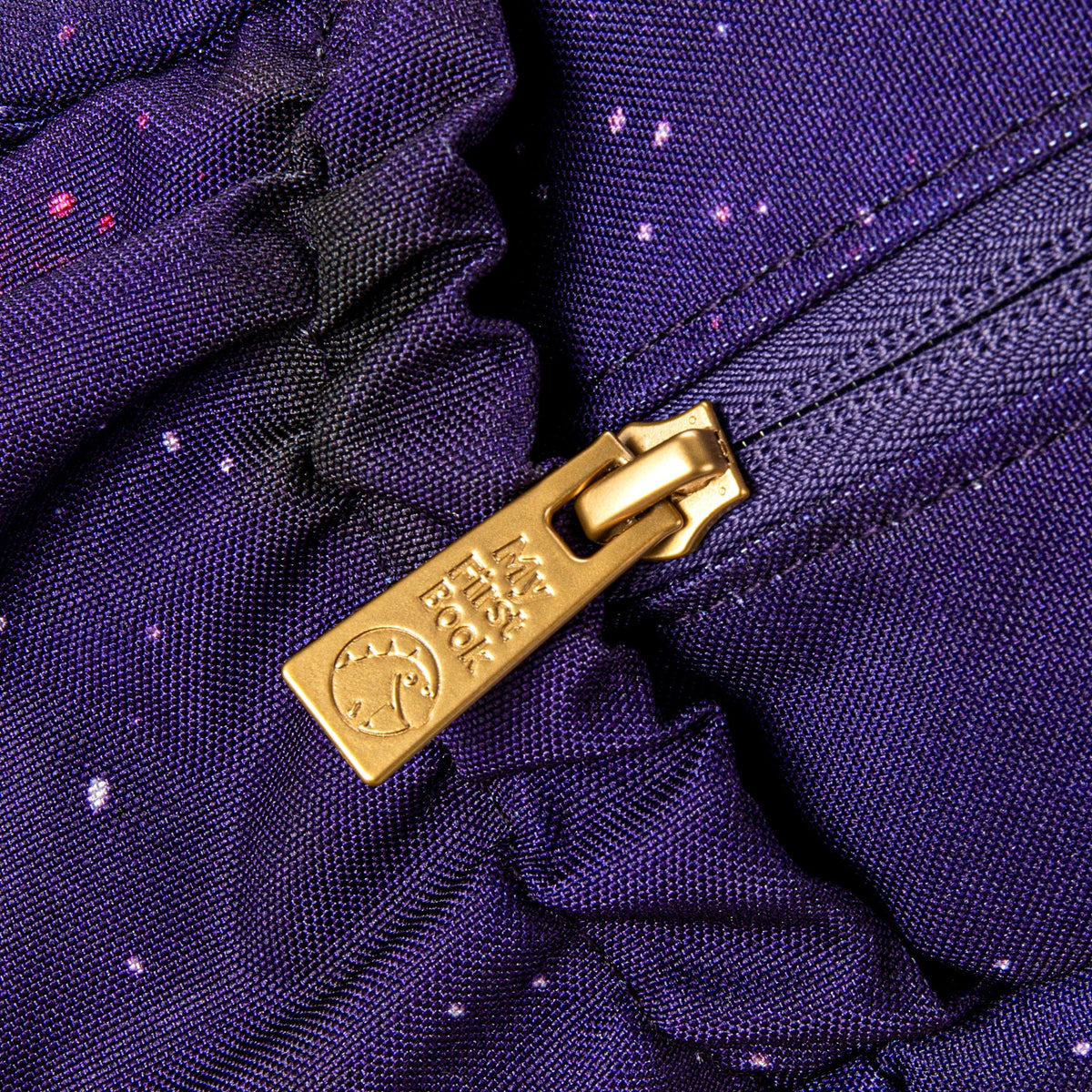Close-up of the golden zip with the -My First Book- logo on the purple galaxy space backpack