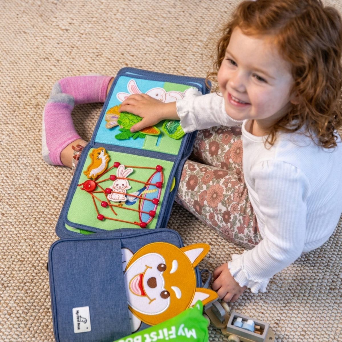 Smiling girl lies on the rug playing with the rabbit meadow page and the birdcage page of the Pet Friends quiet book, the matching Shiba dog backpack in the foreground. From 1 year.
