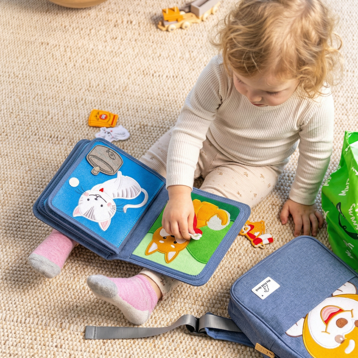 Play scene: toddler sits on the rug and plays with focus on the open dog and cat page of the Pet Friends quiet book, the matching dog backpack beside it. From 1 year.