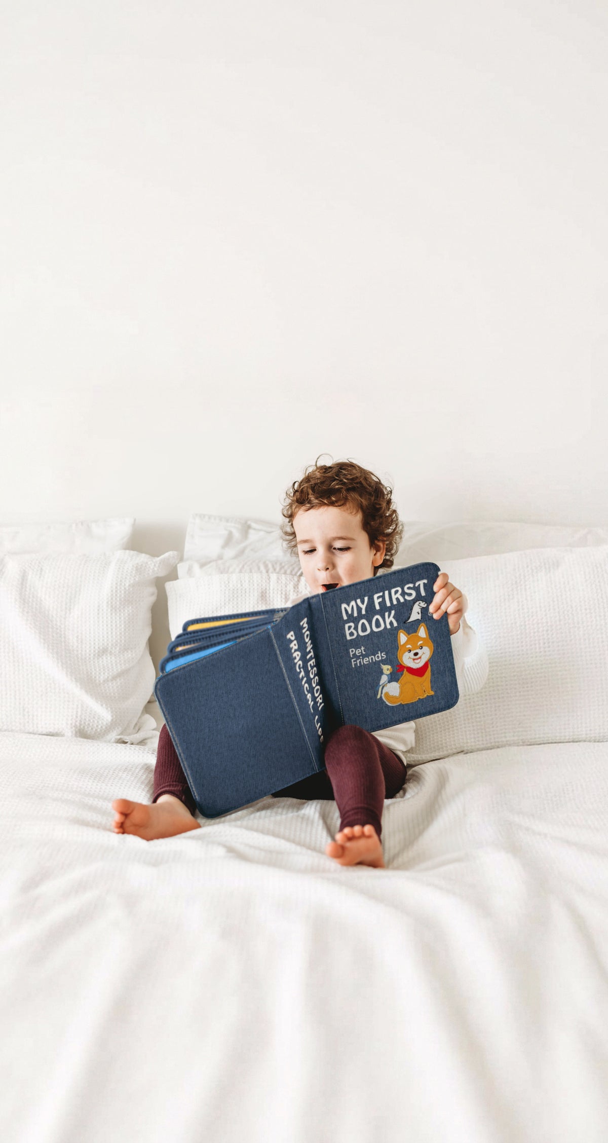 Kleinkind sitzt auf einem weißen Bett und liest im My First Book Pet Friends Quiet Book